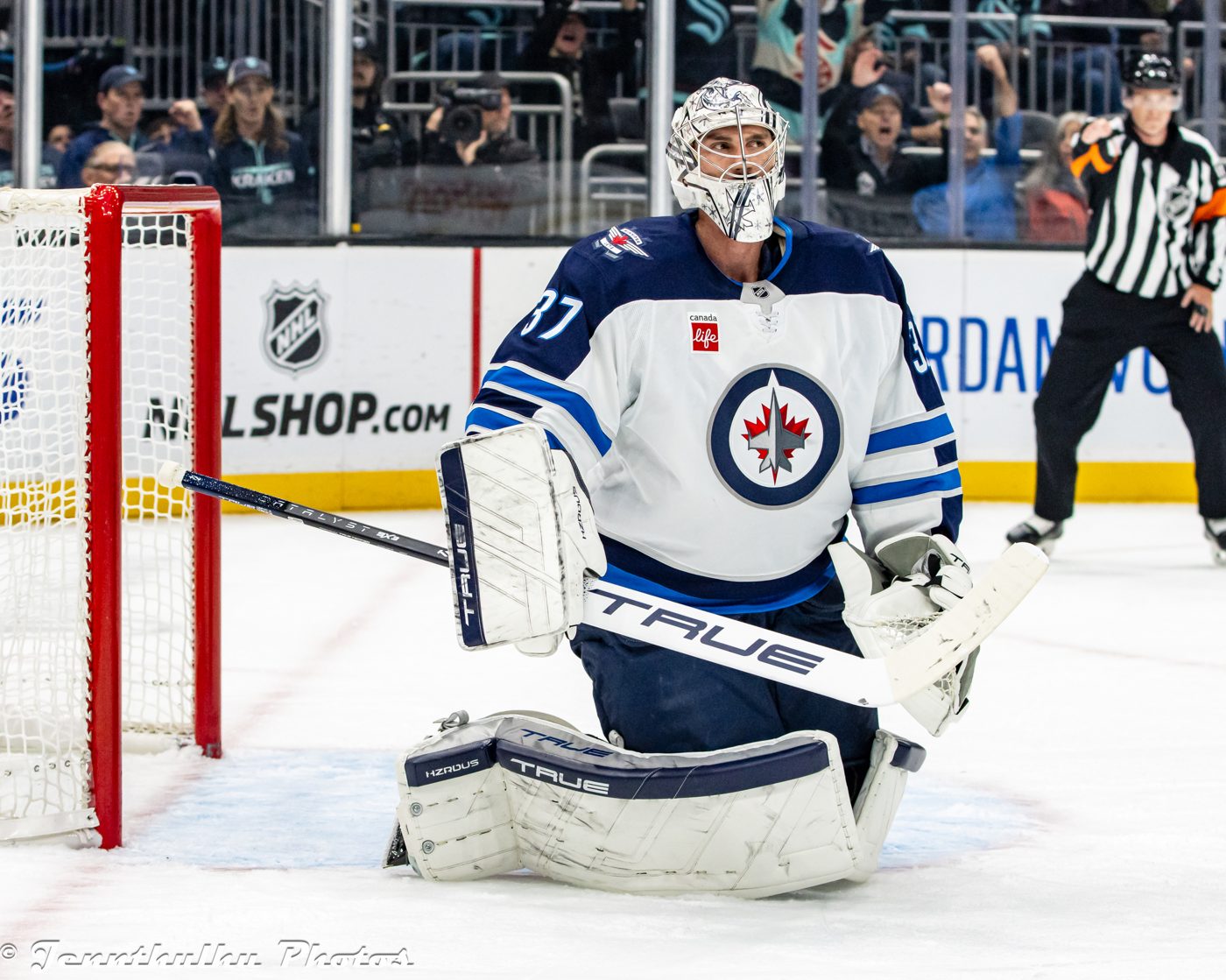 Connor Hellebuyck in a white Winnipeg Jets jersey, kneeling in his crease and looking up the ice.